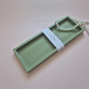 Peut inclure: Un plateau rectangulaire vert sauge avec une bordure surélevée, enveloppé d'une bande blanche avec le texte "The Sage Rose Studio". Un rang de perles blanches repose sur le plateau, sur fond rose.