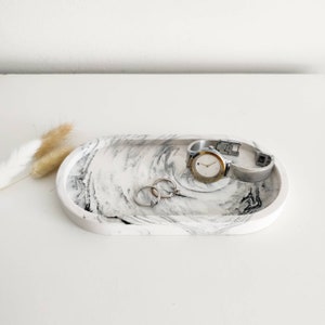May include: White oval tray with black marbling, featuring a silver watch and two silver rings.