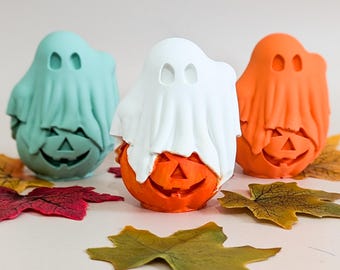 May include: Three Halloween decorations: a teal ghost, a white ghost, and an orange ghost, each atop a carved pumpkin. The pumpkins have jack-o'-lantern faces. Colourful autumn leaves are scattered around the decorations.