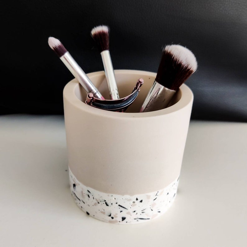A beige and white terrazzo makeup brush holder with three makeup brushes inside. The holder has a smooth, light beige top and a bottom half with a white terrazzo pattern.