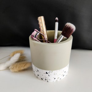 May include: A light gray ceramic makeup holder with a white and black speckled design. The holder contains a pink eyelash curler, a pink and gold lipstick, a pink lip liner, and a brown makeup brush.