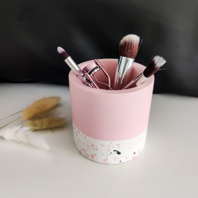 A pink and white terrazzo makeup brush holder with three makeup brushes and a lash curler inside.