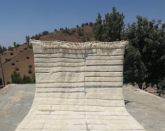 May include: A white and blue striped rug with a fringe border. The rug is folded in half and is lying on a concrete surface. The rug is in a natural setting with a hillside in the background.