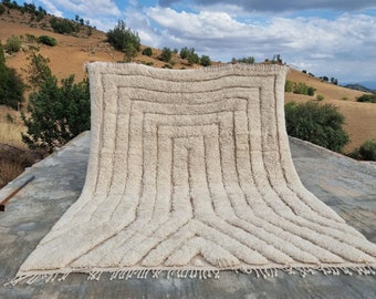 May include: A white, hand-woven rug with a geometric pattern. The rug has a fringe border and is laid out on a concrete surface.