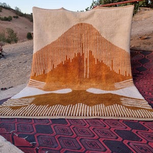 May include: A rectangular area rug featuring a cream-colored background with a large, central mountain-like graphic in shades of burnt orange and brown. The design incorporates vertical line patterns and solid color blocks.