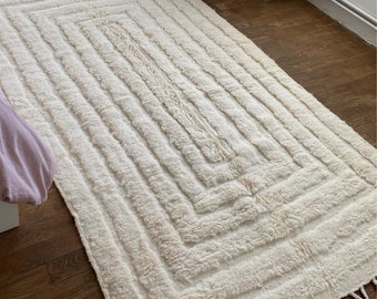 May include: A white, fluffy rug with a geometric pattern. The rug has a fringe border and is made of a soft, textured material.