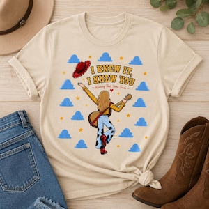 May include: A beige t-shirt featuring a graphic of a person with long hair wearing a yellow plaid shirt and cow-print pants, holding a guitar. The text reads I KNEW IT, I KNEW YOU Wearing That Same Smile, surrounded by blue clouds and yellow stars.