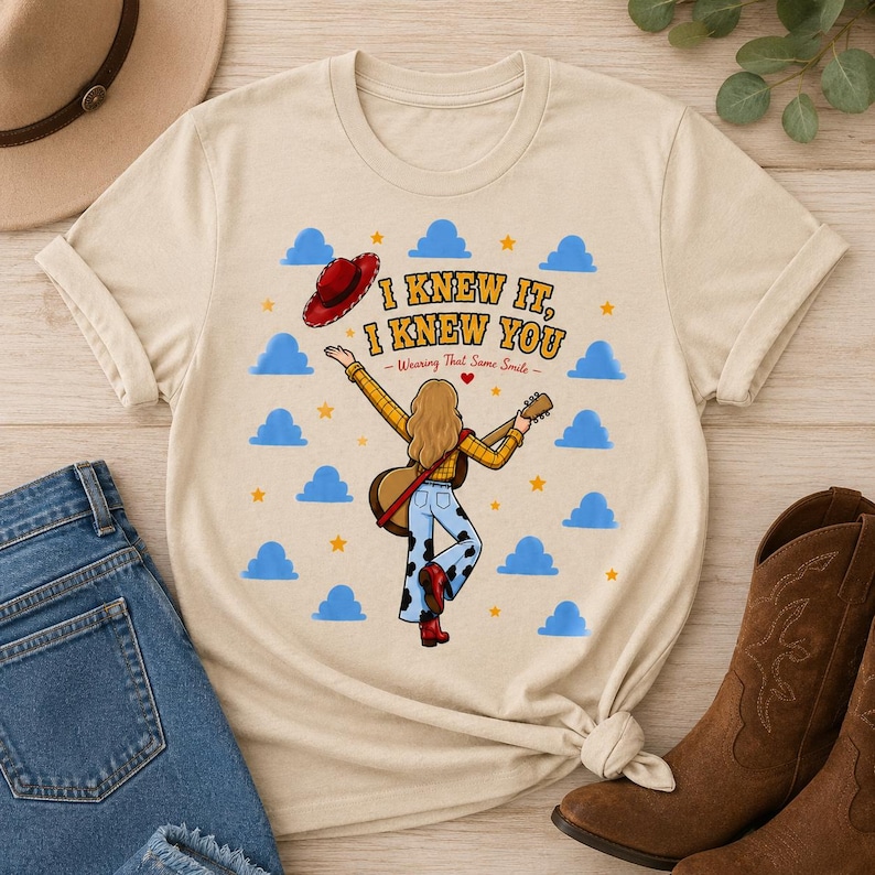 May include: A beige t-shirt featuring a graphic of a person with long hair wearing a yellow plaid shirt and cow-print pants, holding a guitar. The text reads I KNEW IT, I KNEW YOU Wearing That Same Smile, surrounded by blue clouds and yellow stars.