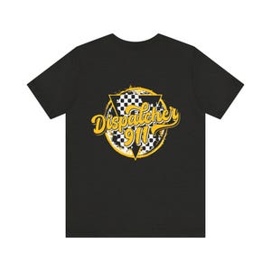 May include: A black short-sleeve t-shirt featuring a graphic design on the chest. The design includes a yellow circle with a distressed edge, a black and white checkered triangle, and the yellow text "Dispatcher 911" in a cursive font.