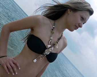 May include: A woman with long brown hair wearing a black bikini top and a seashell necklace. The necklace is made of various seashells, including starfish, and is strung on a brown cord.