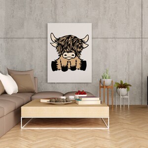 May include: A white canvas print with a cartoon illustration of a brown and black Highland cow sitting with its legs crossed. The print is hanging on a gray concrete wall in a living room setting.