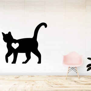 May include: A black silhouette of a cat with a white heart on its chest. The cat is walking to the right. The image is on a white wall with a pink chair and a potted plant in the background.
