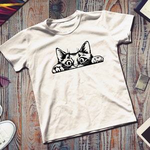 May include: White t-shirt with a black and white graphic of a cat peeking over the edge of a surface.