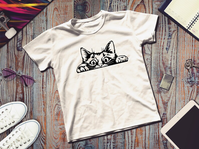 May include: White t-shirt with a black and white graphic of a cat peeking over the edge of a surface.