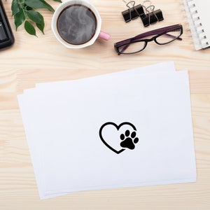 May include: A black and white paw print inside a heart shape on a white sheet of paper. The paper is on a light wood surface.