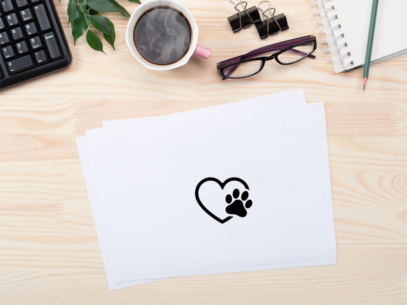 May include: A black and white paw print inside a heart shape on a white sheet of paper. The paper is on a light wood surface.