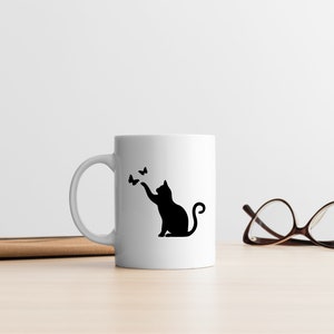 May include: White ceramic mug with a black silhouette of a cat reaching for butterflies. The mug is on a wooden surface next to a pair of brown eyeglasses.