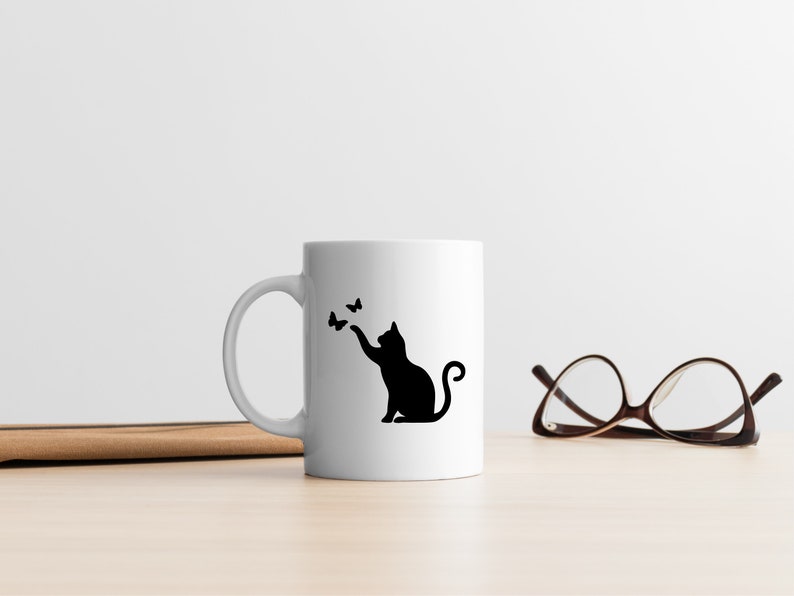 May include: White ceramic mug with a black silhouette of a cat reaching for butterflies. The mug is on a wooden surface next to a pair of brown eyeglasses.
