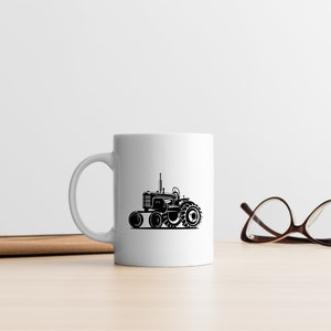 Op de afbeelding: Een witte keramische mok met een zwart silhouet van een tractor aan de voorkant. De mok staat op een houten tafel naast een paar bruine brillen.
