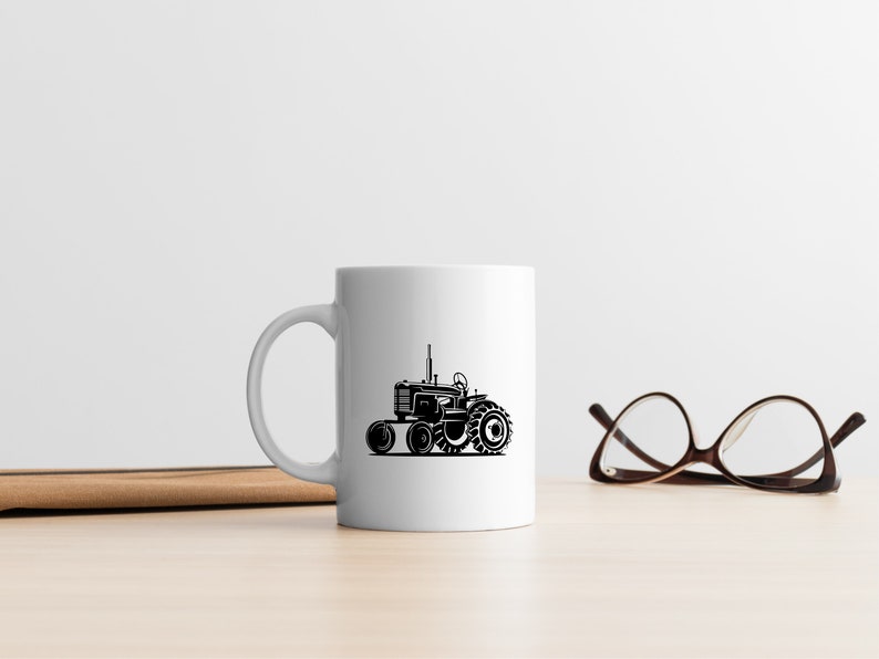 Op de afbeelding: Een witte keramische mok met een zwart silhouet van een tractor aan de voorkant. De mok staat op een houten tafel naast een paar bruine brillen.