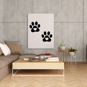 May include: A white canvas print with two black paw prints with white hearts in the center of each print. The print is hanging on a gray concrete wall above a brown sectional sofa with pillows. A wooden coffee table with books, a bowl of decorative items, and a bowl of red berries is in front of the sofa.