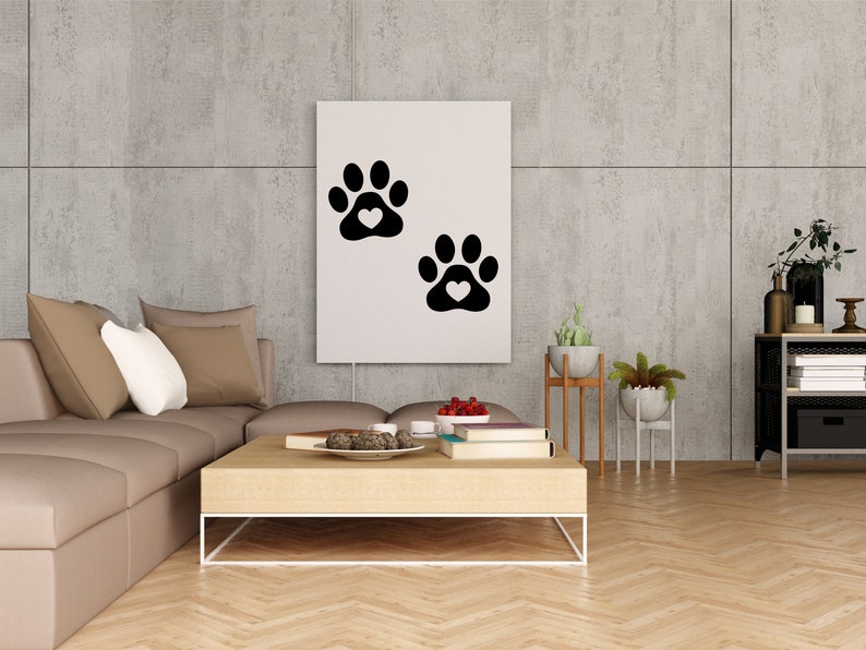 May include: A white canvas print with two black paw prints with white hearts in the center of each print. The print is hanging on a gray concrete wall above a brown sectional sofa with pillows. A wooden coffee table with books, a bowl of decorative items, and a bowl of red berries is in front of the sofa.