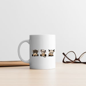 May include: White ceramic mug with a design of three cartoon Highland cows in different poses. The cows are brown and white with black horns and hooves.