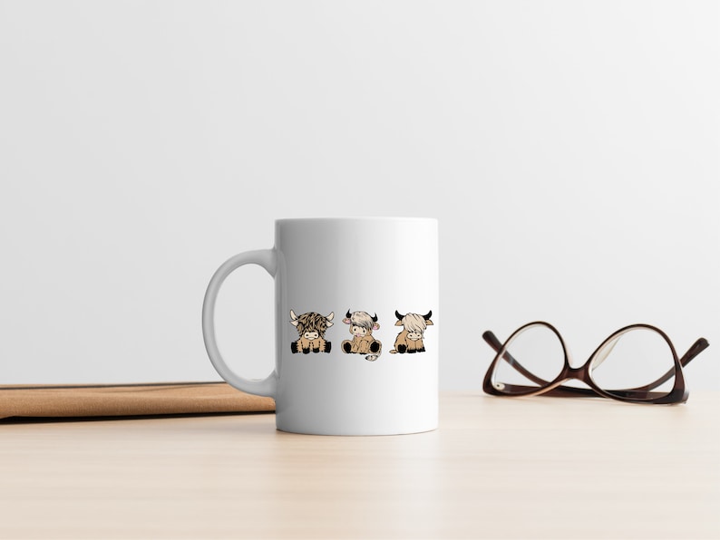 May include: White ceramic mug with a design of three cartoon Highland cows in different poses. The cows are brown and white with black horns and hooves.