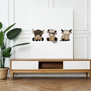May include: Three cartoon Highland cows, each with a different expression, are depicted in a white frame. The cows are brown and white with fluffy fur. The frame is on a white wall above a wooden shelf.