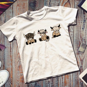 May include: White t-shirt with a graphic of three cartoon Highland cows. The cows are brown and white and have black eyes and noses. They are sitting down and looking to the right.