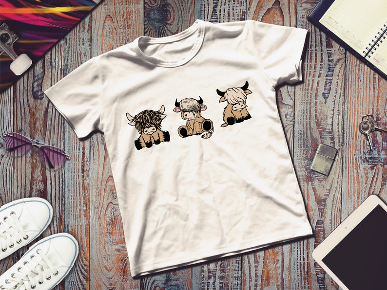 May include: White t-shirt with a graphic of three cartoon Highland cows. The cows are brown and white and have black eyes and noses. They are sitting down and looking to the right.