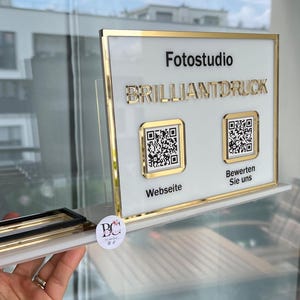 Może przedstawiać: Biały i złoty szyld z napisem "Fotostudio BRILLANTDRUCK" i dwoma kodami QR. Szyld jest trzymany w dłoni i ma złotą ramę. Na szyldzie znajdują się również napisy "Webseite" i "Bewerten Sie uns".