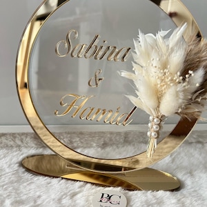 Puede incluir: Un soporte redondo de espejo dorado con un círculo acrílico transparente en la parte superior. El círculo acrílico tiene los nombres "Sabrina & Hamid" escritos en cursiva. Un arreglo de flores secas está unido al círculo acrílico.