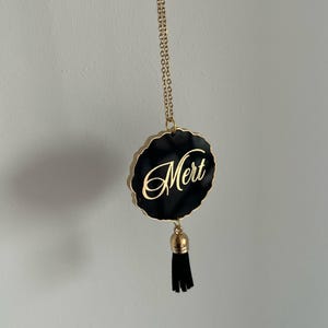 Puede incluir: Un collar de cadena dorada con un colgante negro que presenta el nombre "Mert" en letras doradas. El colgante tiene una borla negra.
