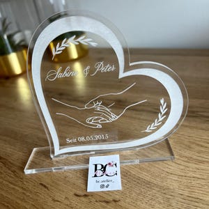 May include: Clear acrylic heart-shaped wedding keepsake with the names "Sabine & Peter" and the date "08.05.2015" etched in white. Features an illustration of two hands clasped together and decorative leaf accents. Includes a small business card.