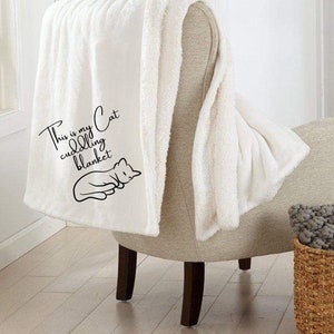 May include: White plush throw blanket with the text "This is my cat cuddling blanket" and a black silhouette of a cat sleeping on it. The blanket is draped over a beige armchair with a gray knitted throw pillow in a wicker basket.