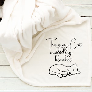 May include: Cream-colored cat cuddling blanket with black text that reads "This is my Cat cuddling blanket" and a black cat illustration. The blanket is folded and draped over a white wooden surface.