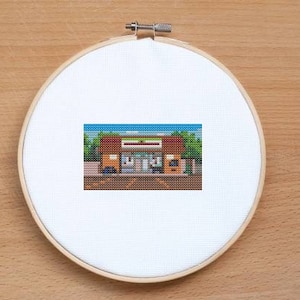 May include: Cross-stitch pattern of a convenience store with a brown brick facade, a green awning, and a blue sky. The store has a sign that reads "7-Eleven".