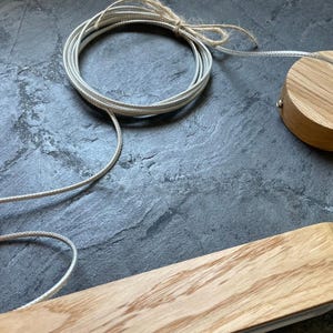 May include: Close-up of wooden light fixture components. The image shows a circular ceiling mount, a rectangular light fixture, and a coiled cord. The wood has a natural grain, and the cord is a light grey colour. The components are on a dark grey surface.