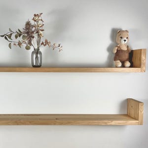 Peut inclure: Deux étagères horizontales en bois brun clair avec des extrémités en forme de U fixées au mur. L'étagère supérieure contient un bocal en verre transparent avec des fleurs séchées, un vase en verre gris avec du feuillage et un petit ours en peluche.