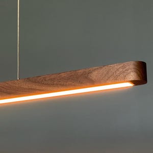 Może przedstawiać: Liniowa lampa wisząca wykonana z ciemnobrązowego drewna o widocznym usłojeniu. Oprawa ma długi, wąski, prostokątny kształt zawieszony na dw&oacute;ch cienkich przewodach, emitujący ciepłe, jasne światło z dolnej części urządzenia.