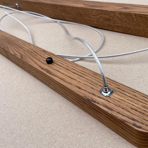 Puede incluir: Una l&aacute;mpara lineal que consta de dos barras de madera paralelas y alargadas con una veta visible. Las barras est&aacute;n conectadas por cables met&aacute;licos trenzados de color plateado que forman bucles entre ellas, y cada barra incluye peque&ntilde;os botones negros.
