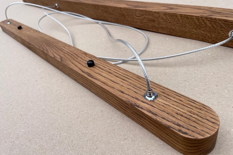 Puede incluir: Una l&aacute;mpara lineal que consta de dos barras de madera paralelas y alargadas con una veta visible. Las barras est&aacute;n conectadas por cables met&aacute;licos trenzados de color plateado que forman bucles entre ellas, y cada barra incluye peque&ntilde;os botones negros.