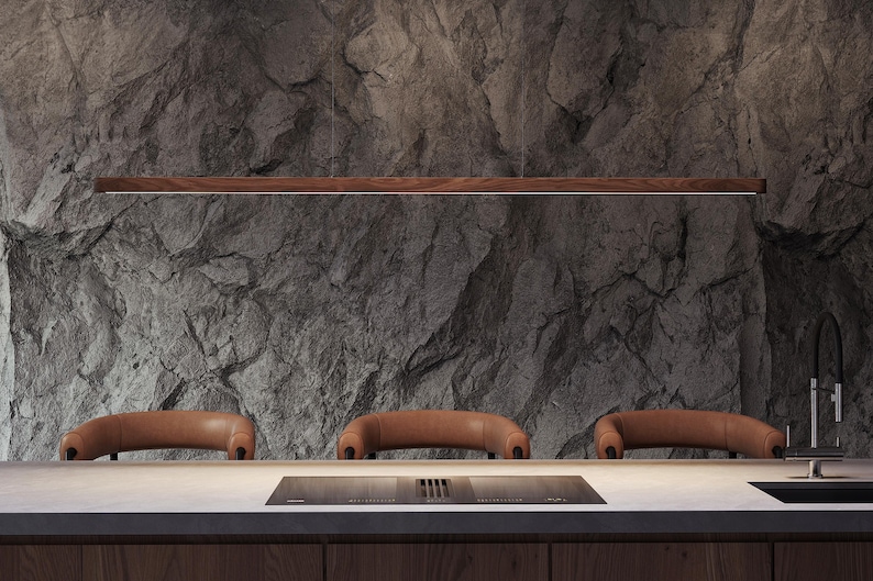 Close-up of a handcrafted black walnut linear pendant light with soft rounded edges, suspended above a modern kitchen island with dark stone walls and leather bar stools. The warm LED glow highlights the natural wood grain and minimalist design.