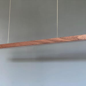 Może przedstawiać: Liniowa lampa wisząca z długą, poziomą drewnianą belką o widocznym ciemnobrązowym usłojeniu. Oprawa jest zawieszona na dw&oacute;ch cienkich, pionowych linkach i posiada biały panel świetlny wzdłuż dolnej krawędzi.