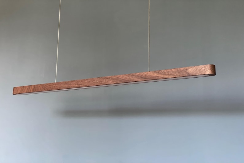 Może przedstawiać: Liniowa lampa wisząca z długą, poziomą drewnianą belką o widocznym ciemnobrązowym usłojeniu. Oprawa jest zawieszona na dw&oacute;ch cienkich, pionowych linkach i posiada biały panel świetlny wzdłuż dolnej krawędzi.
