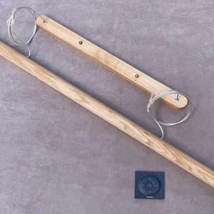 Puede incluir: Una barra de cortina de madera clara, con un panel de control rectangular con un dise&ntilde;o circular. La barra va acompa&ntilde;ada de una pieza de madera m&aacute;s peque&ntilde;a y un cable. Los art&iacute;culos est&aacute;n sobre una superficie gris.