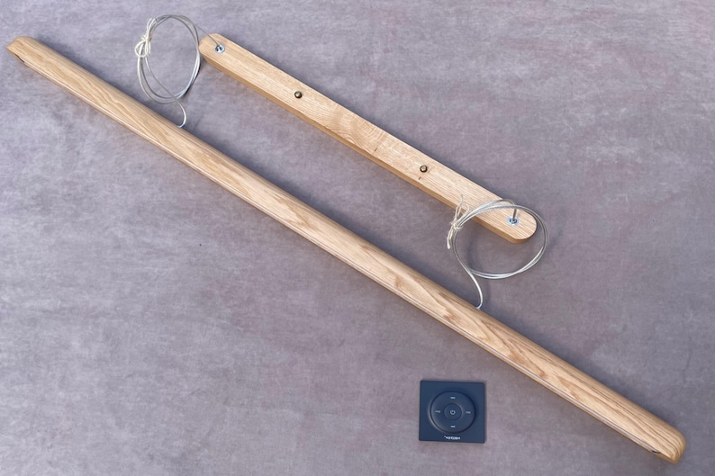 Puede incluir: Una barra de cortina de madera clara, con un panel de control rectangular con un dise&ntilde;o circular. La barra va acompa&ntilde;ada de una pieza de madera m&aacute;s peque&ntilde;a y un cable. Los art&iacute;culos est&aacute;n sobre una superficie gris.