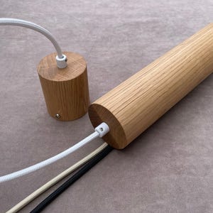Op de afbeelding: Twee houten hanglampen met witte, beige en zwarte snoeren. De ene armatuur is cilindrisch, de andere is een kortere, afgeronde cilinder. Het hout heeft een natuurlijke, lichtbruine kleur.
