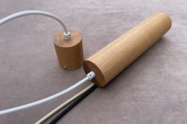 Op de afbeelding: Twee houten hanglampen met witte, beige en zwarte snoeren. De ene armatuur is cilindrisch, de andere is een kortere, afgeronde cilinder. Het hout heeft een natuurlijke, lichtbruine kleur.